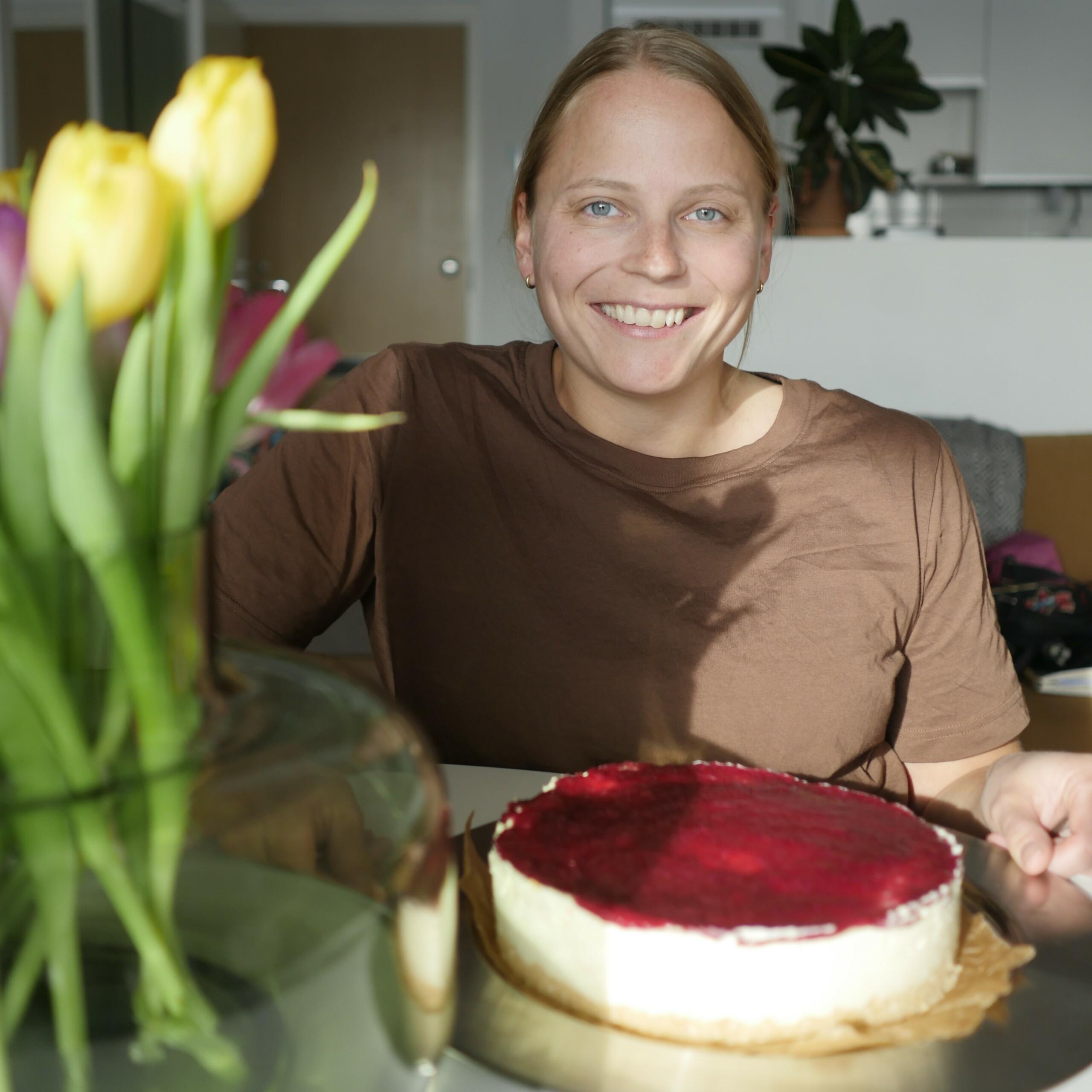 Nainen istuu ruokapöydässä, jossa on juustokakku. Pöydällä on myös keltaisia tulppaaneja.