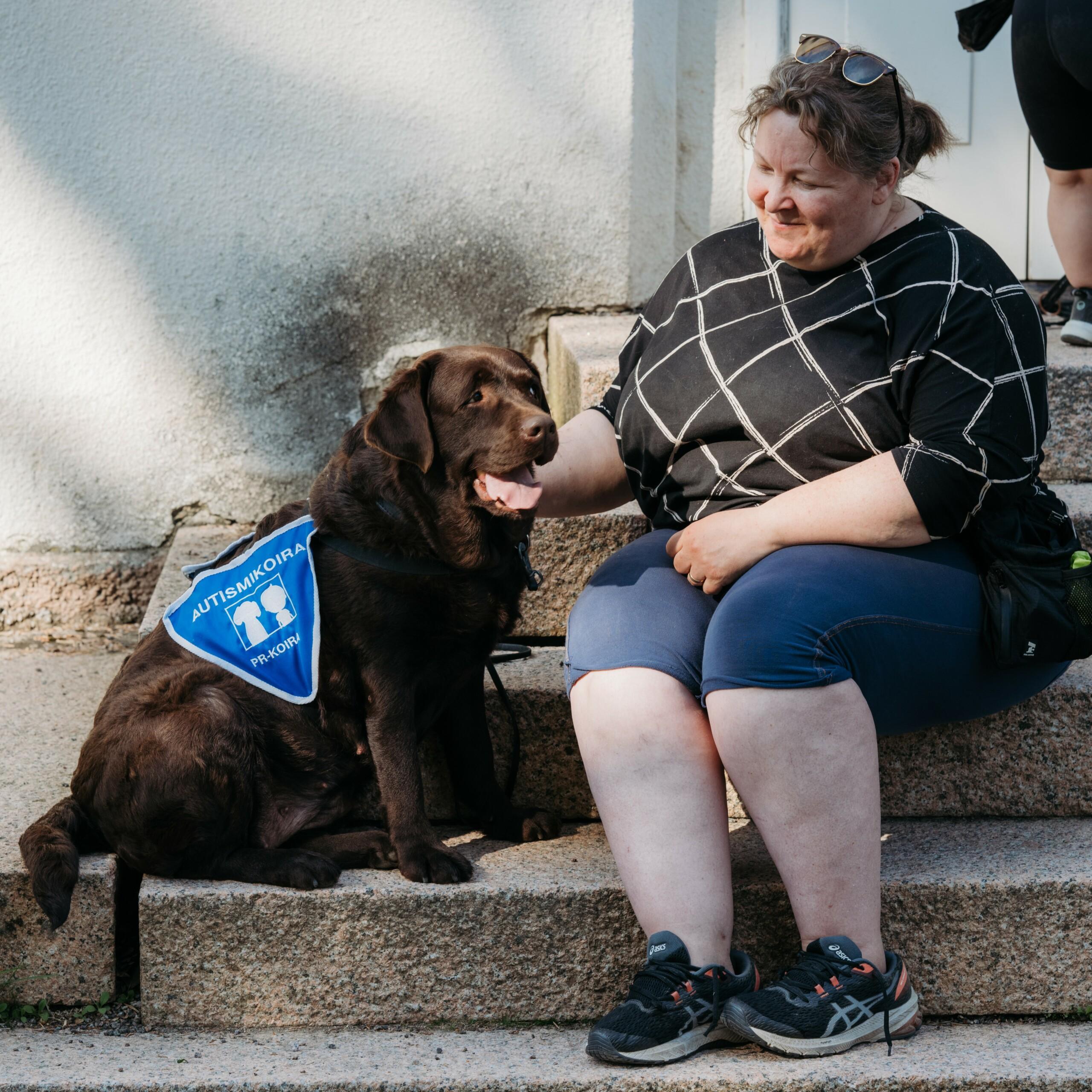 Nainen istuu labradorinnoutajan kanssa rappusilla. Koiralla on päällään sininen autismikoira-liivi.