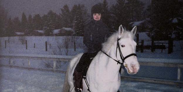 Valkoisen hevosen selässä ratsastaa tummiin pukeutunut nuori nainen. Taustalla on luminen pihamaa.