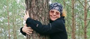 Huivipäinen, tummapuseroinen hymyilevä nainen aurinkolasit päässä, halaa puuta.
