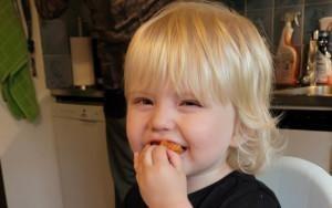 Pieni vaaleatukkainen lapsi laittaa kädellä ruokaa suuhunsa. Hänen ilmeensä on iloinen.