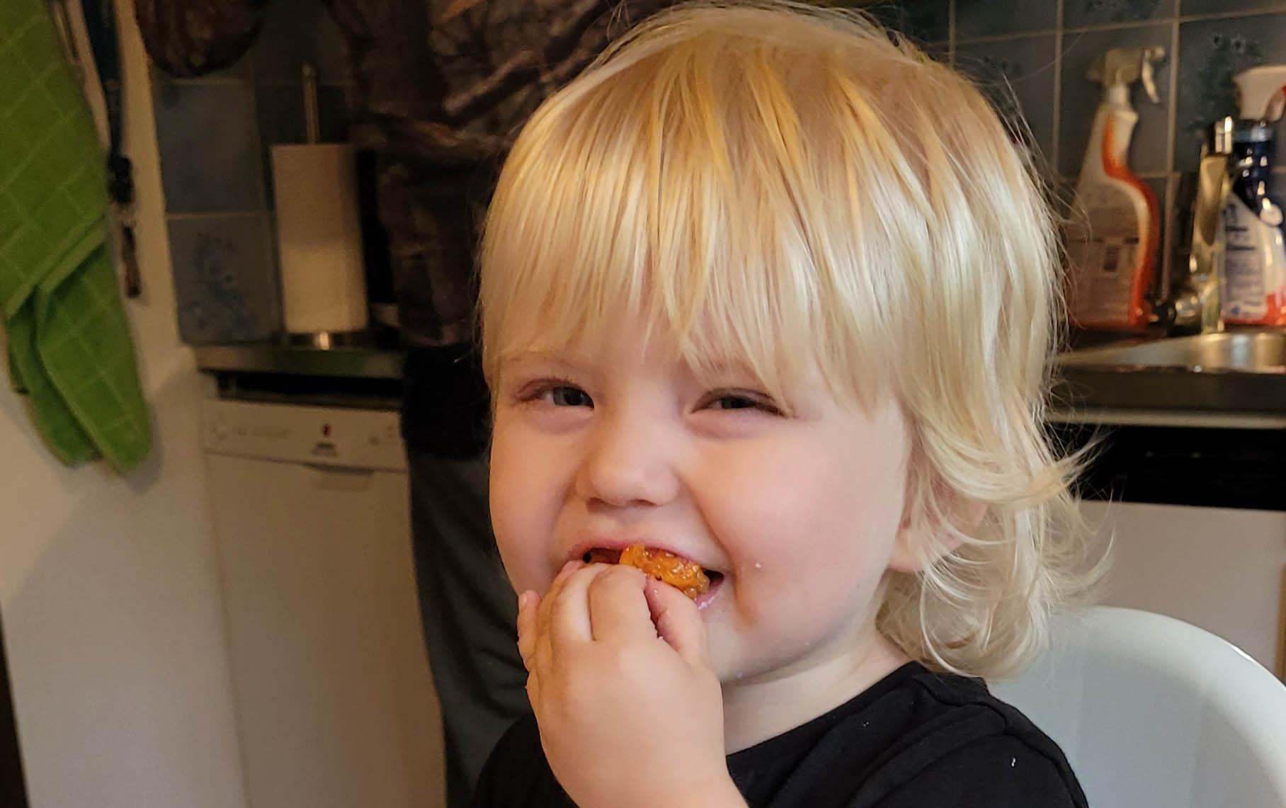Pieni vaaleatukkainen lapsi laittaa kädellä ruokaa suuhunsa. Hänen ilmeensä on iloinen.