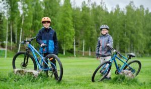 Kaksi poikaa pitelee sinisiä maastopyöriään vihreällä nurmikentällä. Molemmilla on pyöräilykypärät ja he hymyilevät.