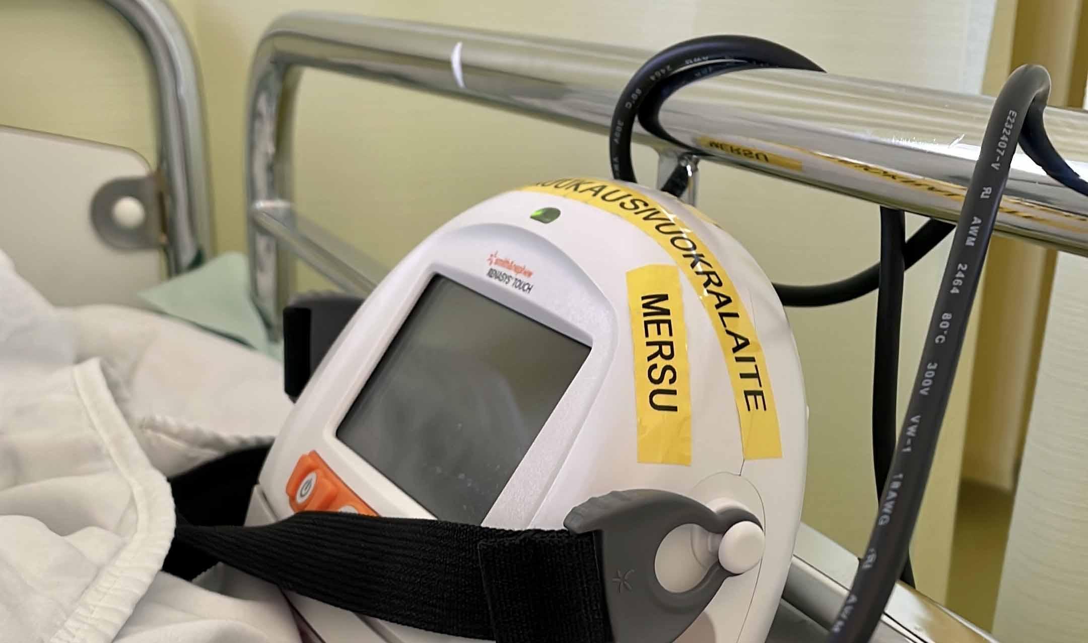 Sairaalasängyllä näkyvä valkoinen, puolipyöreä seurantalaite, jossa keltaisella tarrateipillä teksti MERSU. Takana näkyy sängyn metallipäätyä ja musta johto.