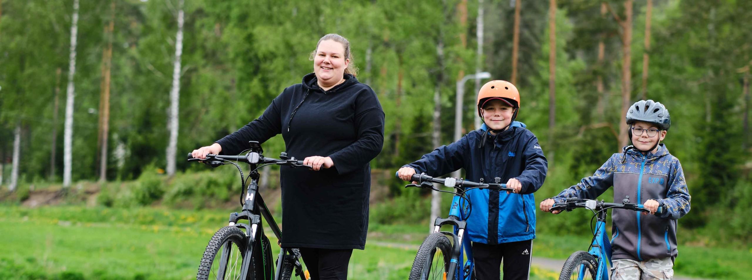 Äiti ja kaksi pientä poikaa taluttavat iloisina uusia maastopyöriään vihreässä kesäisessä maisemassa.
