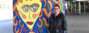 Iloinen, tummatukkainen silmälasipäinen nainen seisoo seinään maalatun iloisen, hymyileväkasvoisen muraalimaalauksen vieressä.